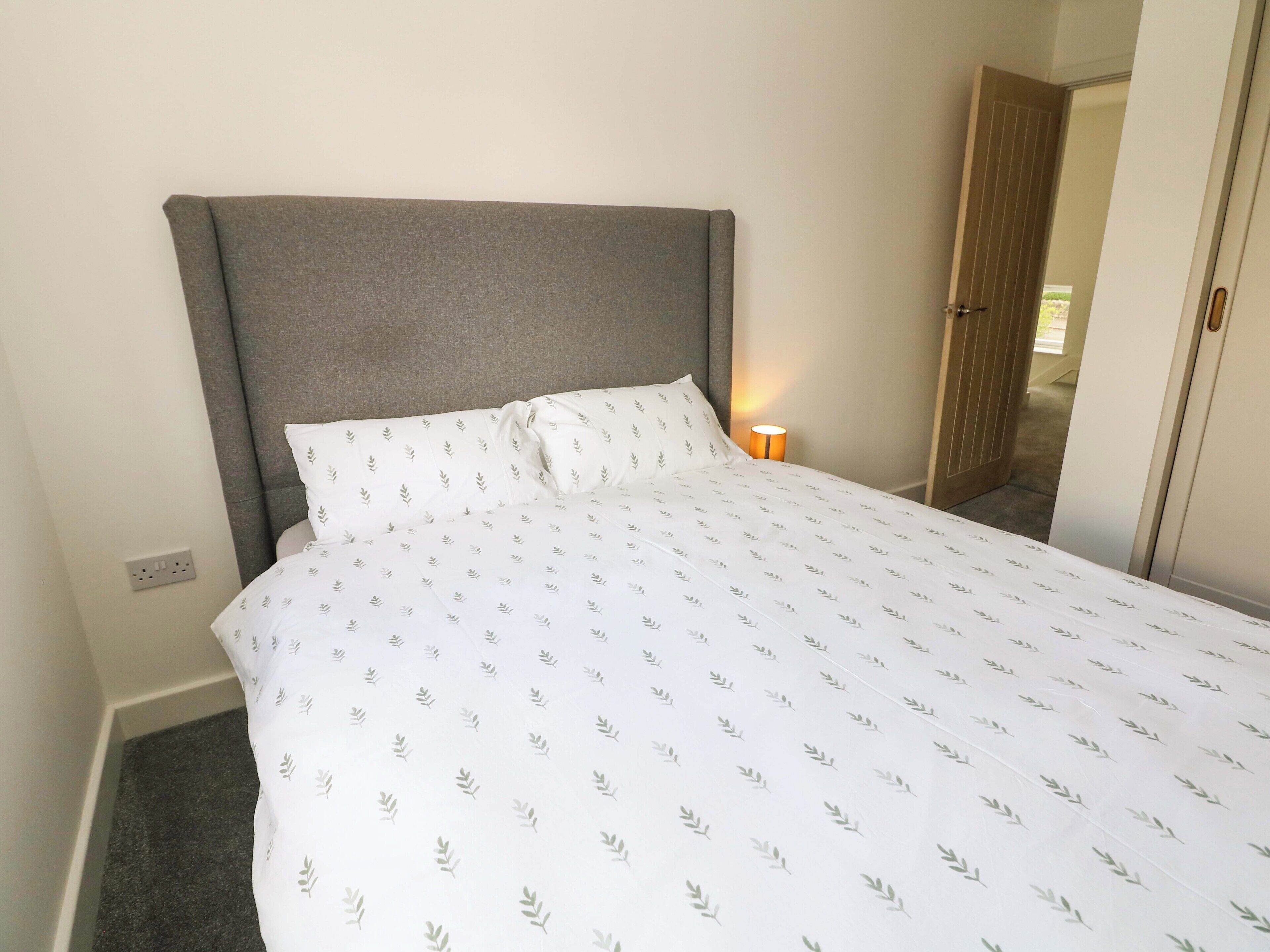 A bed with a grey headboard, white bedding with a leaf pattern, a bedside lamp, and a door to the right.