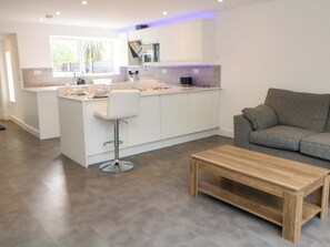 Eine moderne Küche mit weißen Schränken, einem Barhocker und einem grauen Sofa in einem Wohnbereich mit Couchtisch aus Holz.