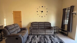 Een woonkamer met een bank, een vloerkleed, een plafondventilator en een wandklok.