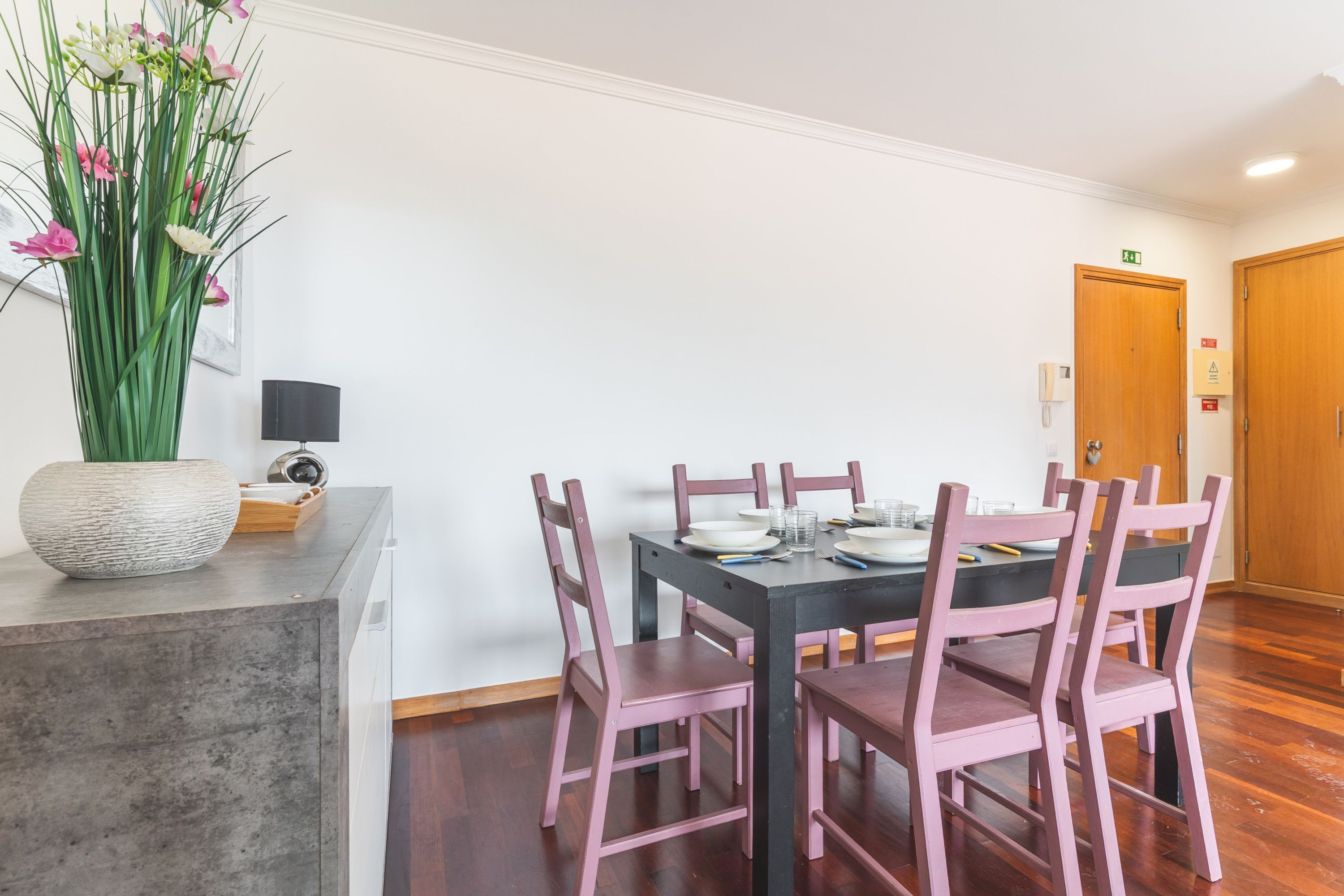 Un coin repas avec une table dressée pour six personnes, entouré de chaises roses, d’un vase rempli de fleurs et d’un luminaire moderne.