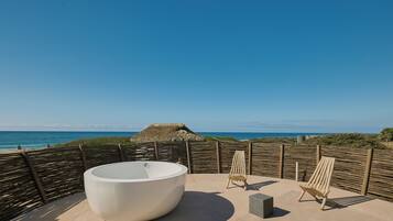 A rooftop with a large bathtub, two lounge chairs, and a view of the ocean.