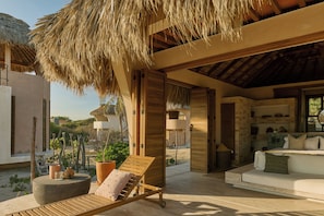 A sunlit outdoor lounge area with a wooden lounge chair, a round table, and a thatched roof structure. - Casa Sal by Tasman (Villa de Tututepec de Melchor Ocampo)