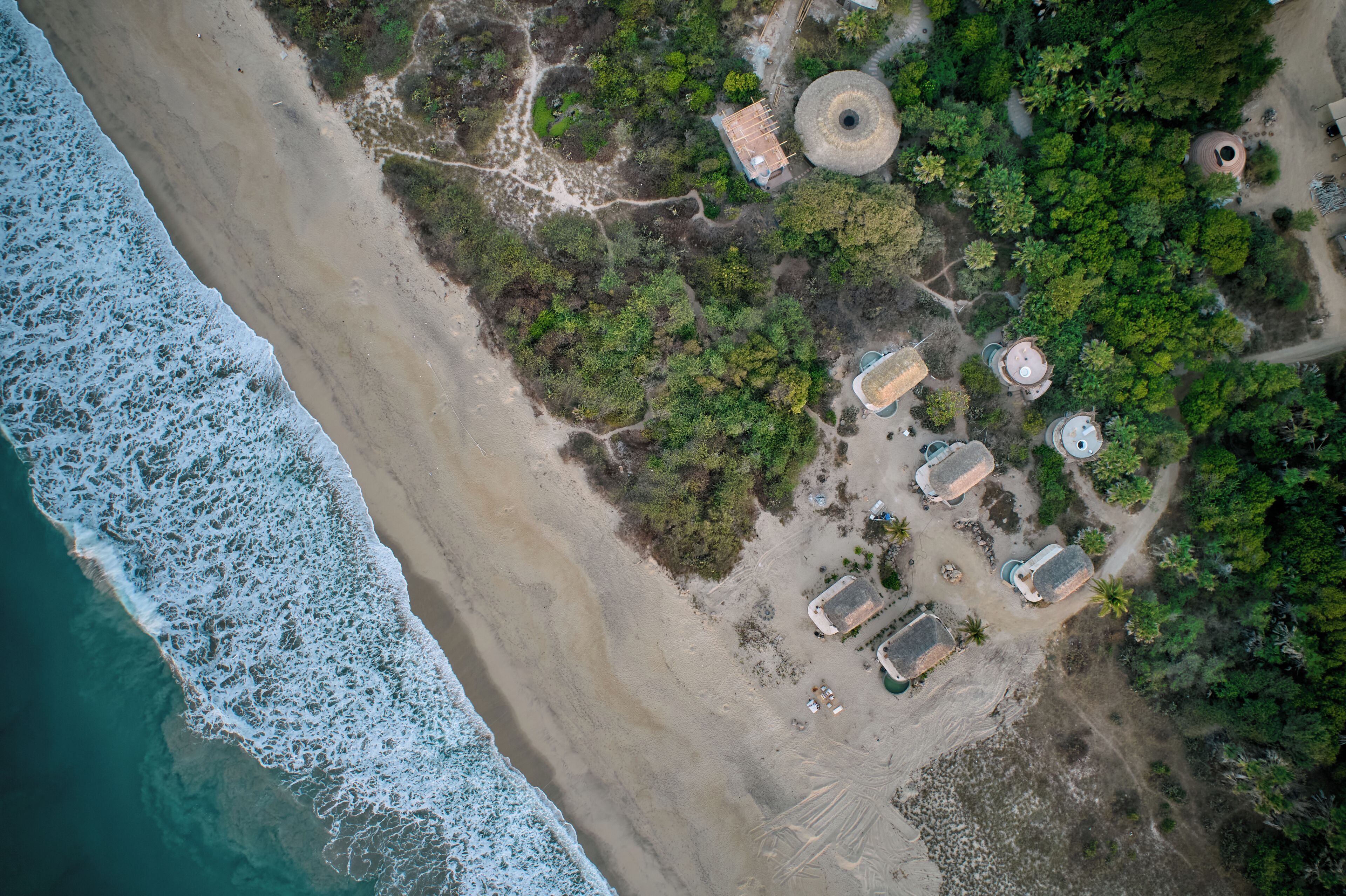 An aerial view of a coastal area with a beach, trees, and several structures with thatched roofs.