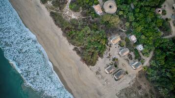 An aerial view of a coastal area with a beach, trees, and several structures with thatched roofs.