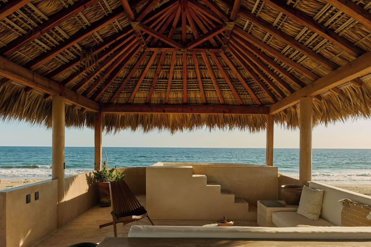Un salón junto a la playa con techo de paja, un sofá, una silla y vistas al océano.