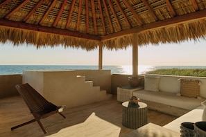 A covered outdoor seating area with a view of the ocean, a wooden chair, a sofa with cushions, and a small table.