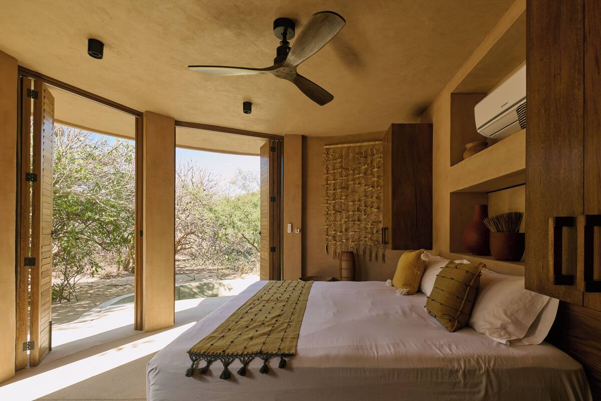 Un dormitorio con una cama grande, un ventilador de techo, una ventana con vista a árboles y un equipo de aire acondicionado instalado en la pared.