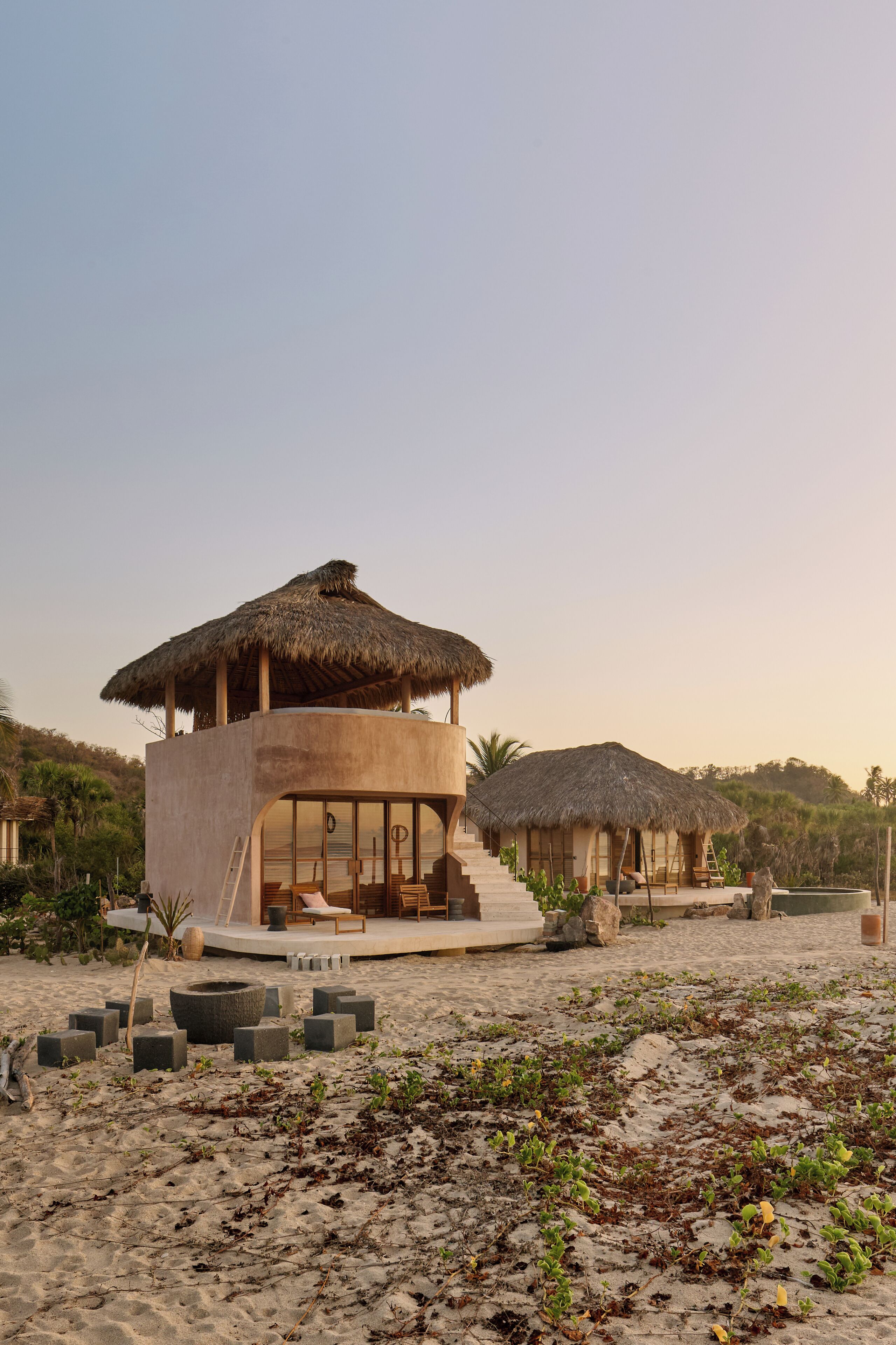 A beachfront property with a central building featuring a thatched roof, a staircase leading to a deck, and a sandy area with scattered tires.