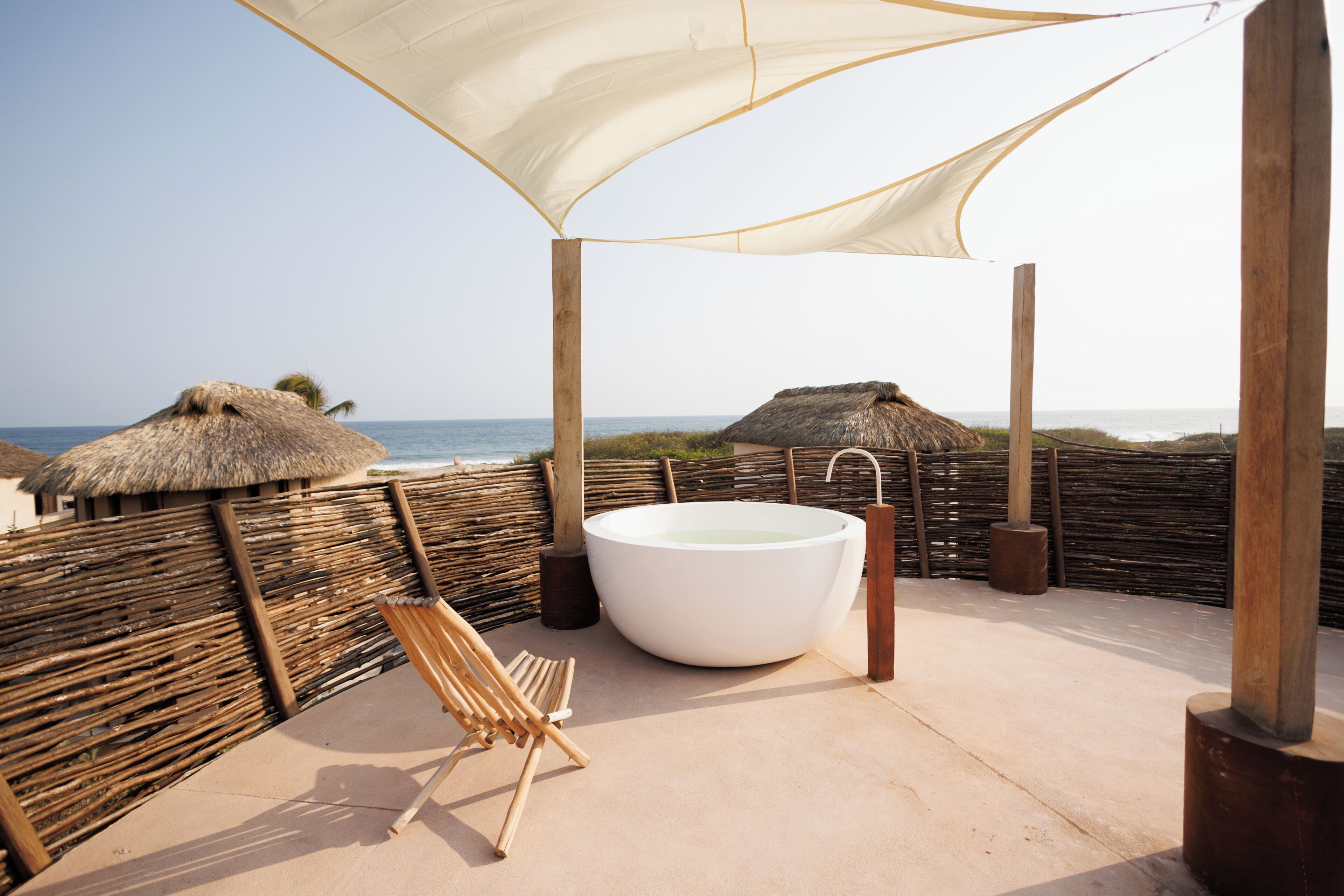 A beachside setting with a large bathtub, a wooden chair, and thatched-roof structures.