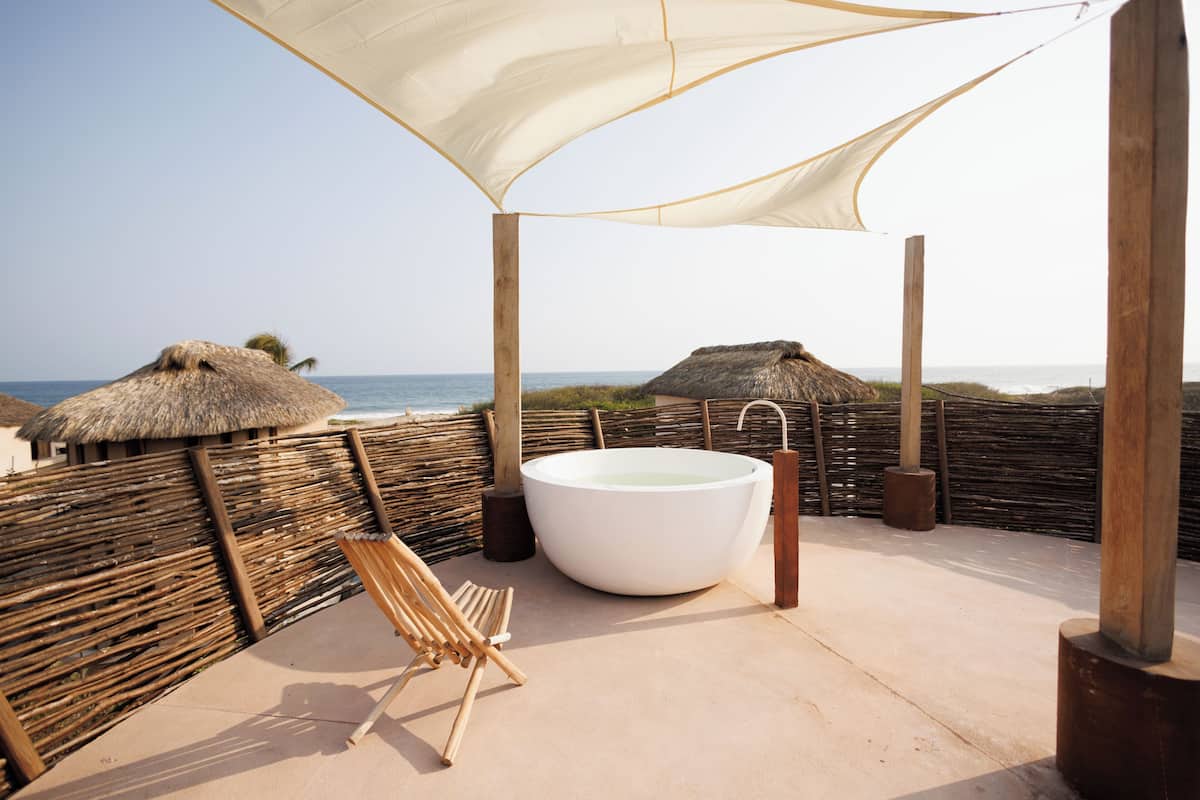 Un paisaje junto a la playa con una bañera grande, una silla de madera y construcciones de techo de paja.