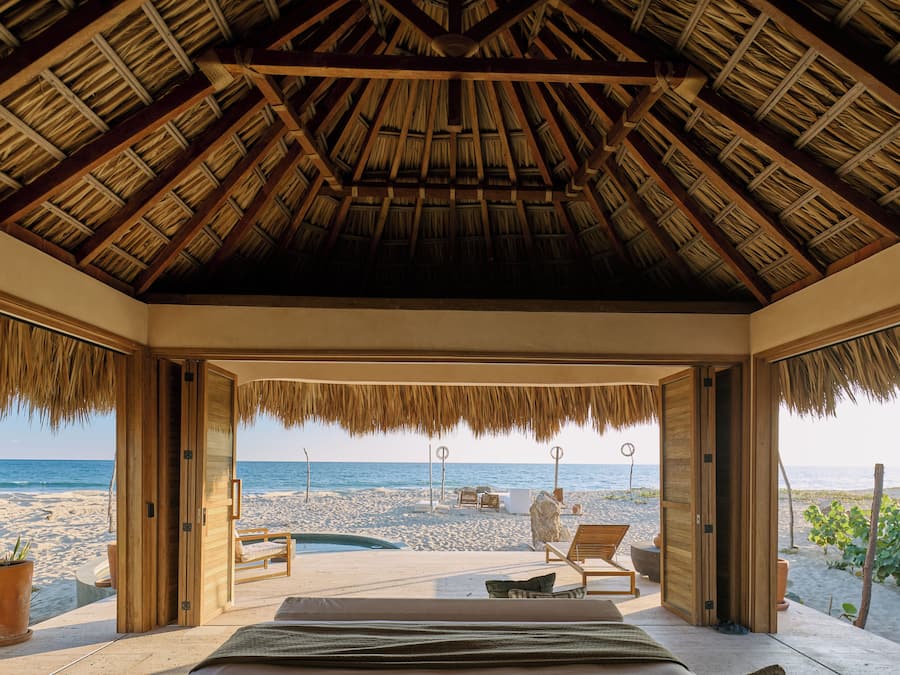 Ein Zimmer am Strand mit einem Reetdach, einem Bett, Blick auf den Strand und einem kleinen Pool.