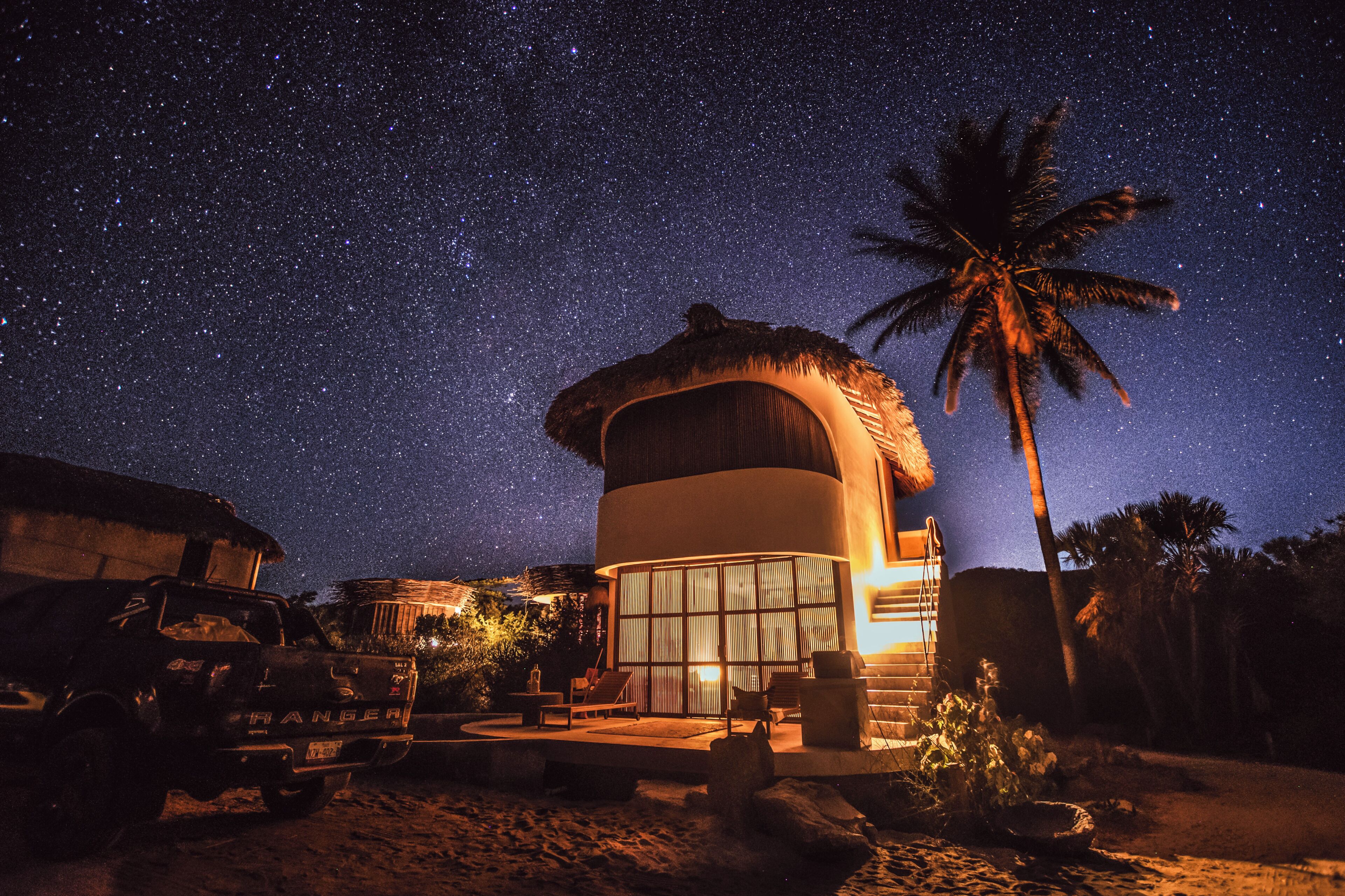A thatched-roof structure with a large window, a palm tree, and a parked vehicle.