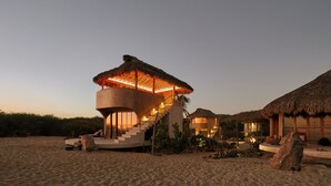 Un resort frente a la playa con una estructura circular, un edificio de techo de paja y una escalera que lleva a un nivel superior.