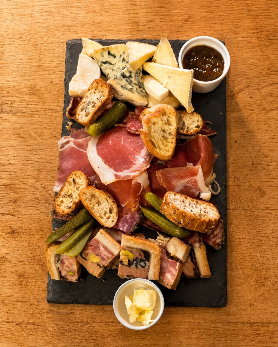 A charcuterie board with various meats, cheeses, pickles, and bread on a slate board.