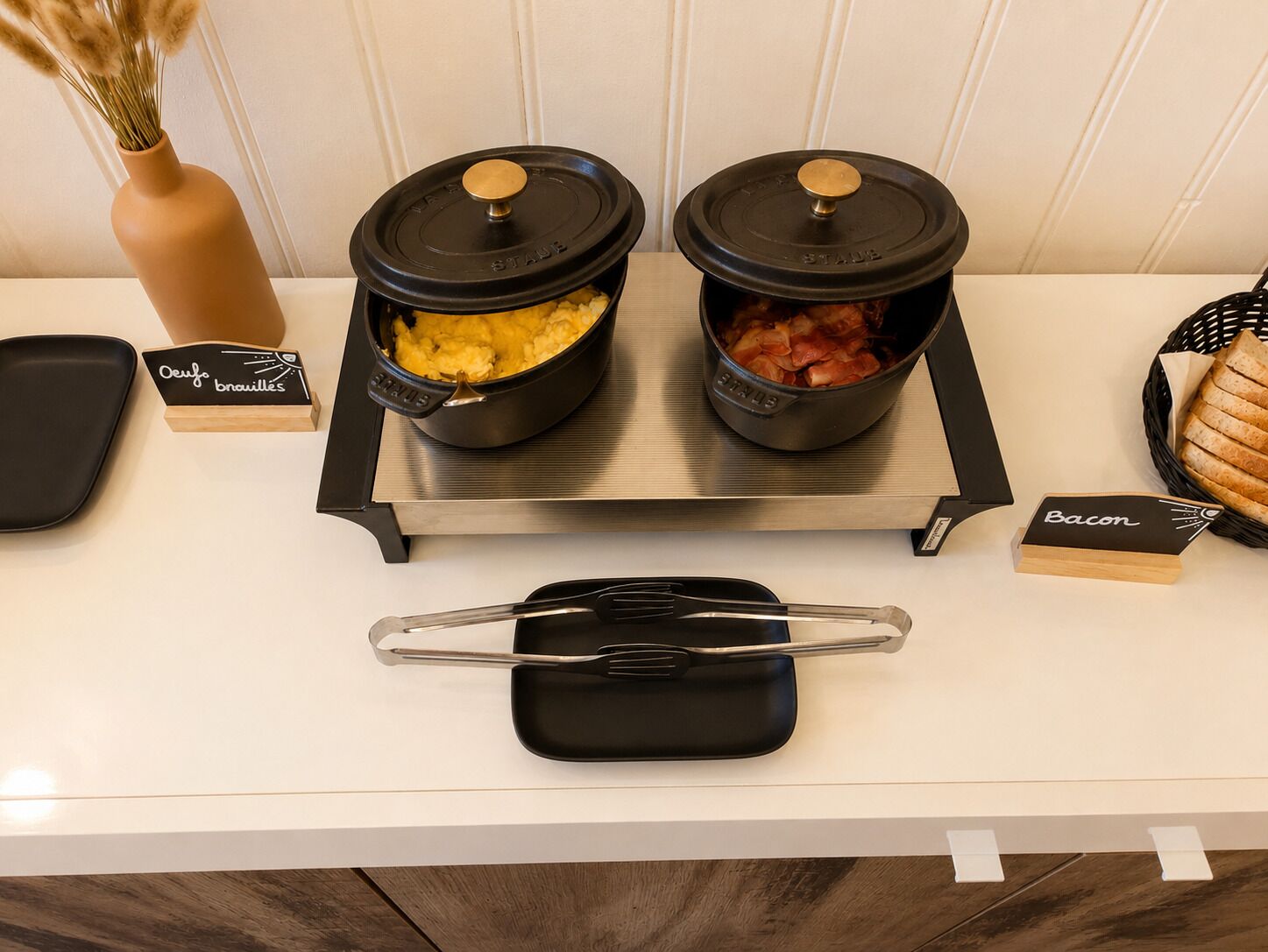 A kitchen counter with two black pots labeled 'Oeuf' and 'Bacon,' a small black pot, and a tray with a spatula.