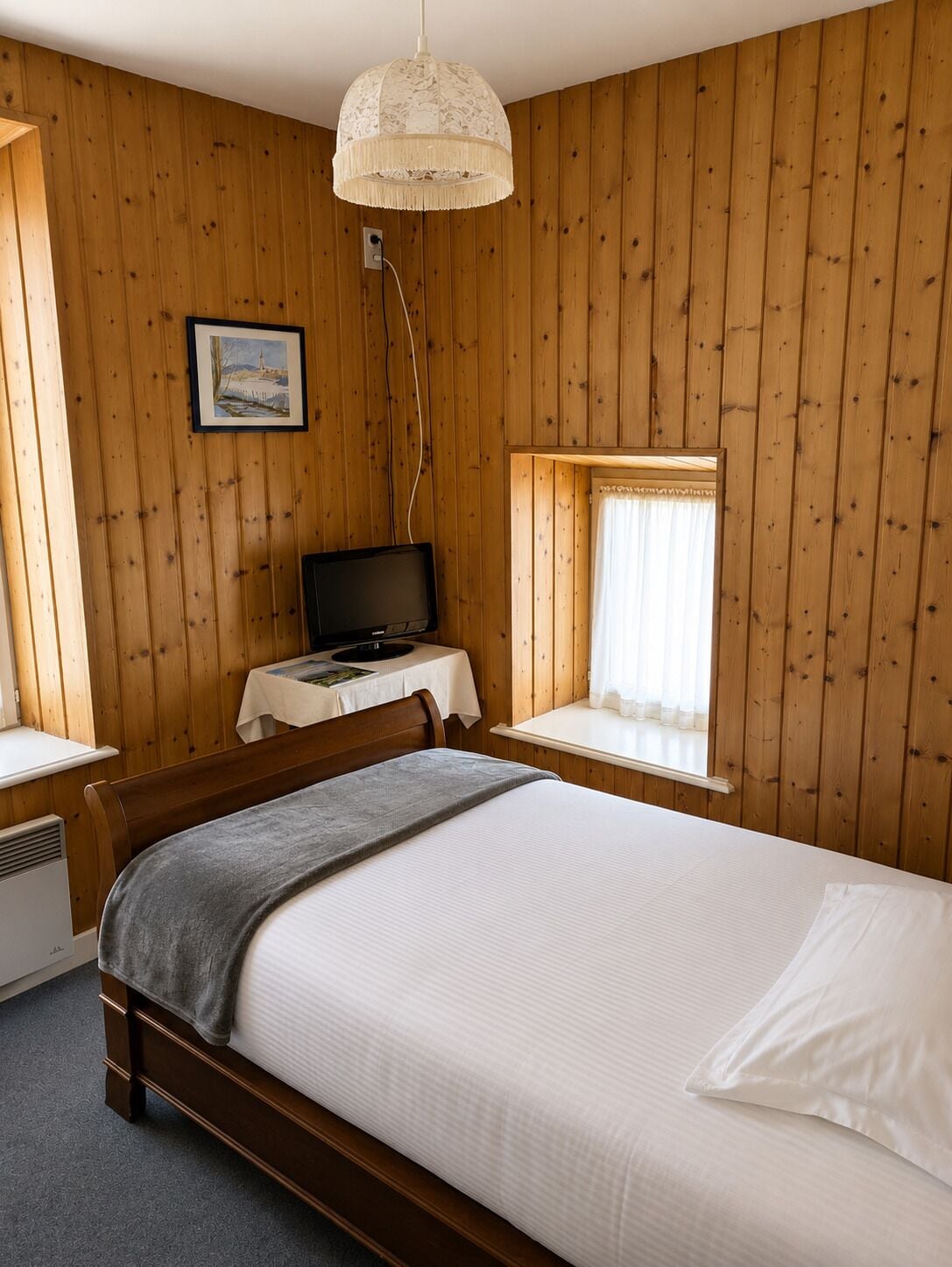 A wooden-paneled room with a bed, a desk, a television, and a window with curtains.
