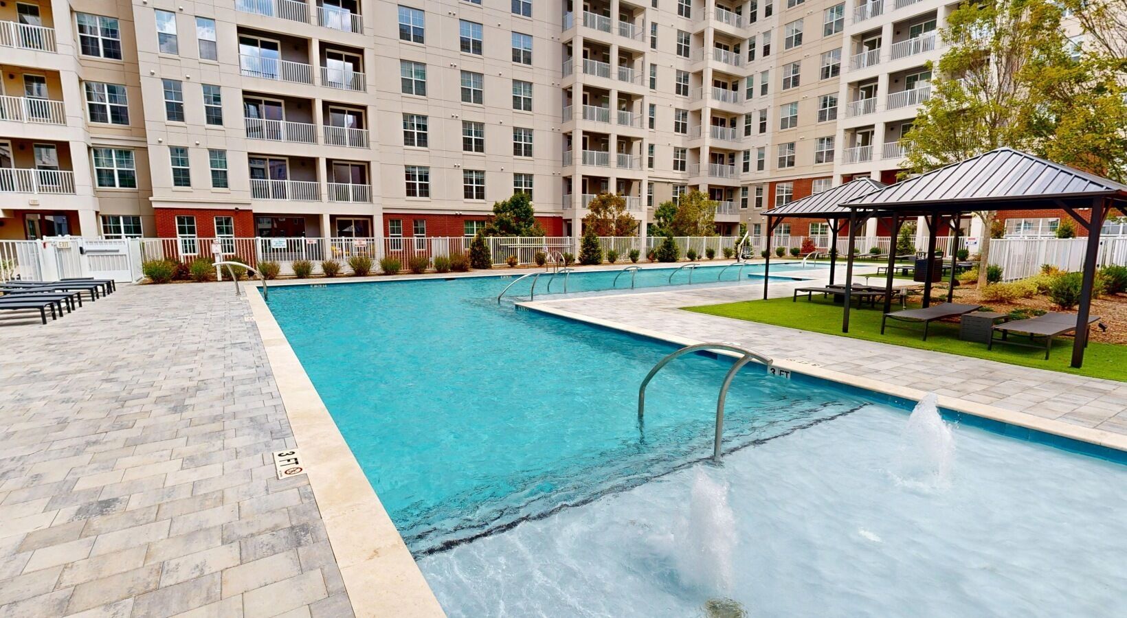 Uma piscina com um deck revestido de azulejos, um gazebo e um edifício de vários andares ao fundo.
