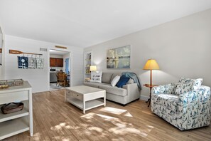 Una sala de estar moderna con un sofá, una mesa de centro y una lámpara. Se observa una cocina al fondo.