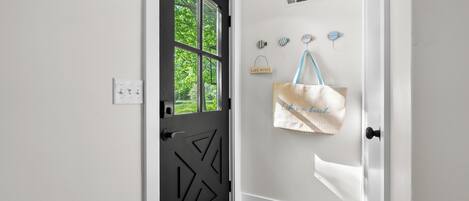 Uma porta preta com padrões geométricos, uma parede branca com uma placa 'LAKE HOUSE' e uma bolsa bege pendurada em um gancho.