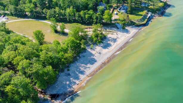 An aerial view of a coastal area with a beach, dense greenery, and a few buildings.