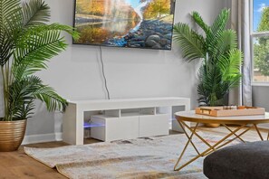 A modern living room with a white entertainment unit, large potted plants, a round coffee table, and a large TV mounted on the wall. - Modern 4BR | Chef`s Kitchen + Soaking Tub · Indy (Indianapolis)