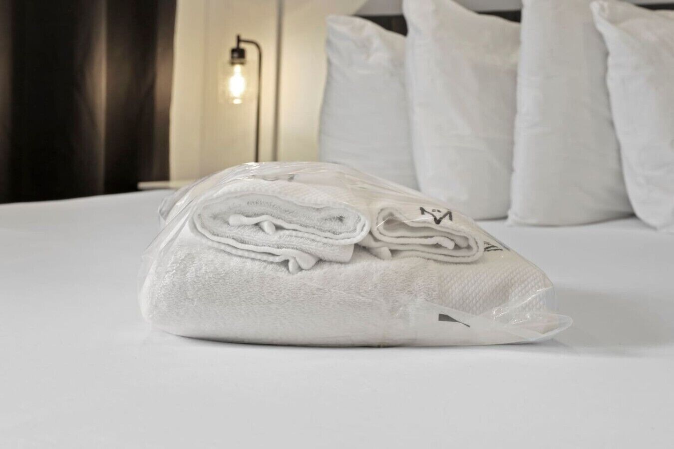 A neatly folded towel on a bed with white bedding, a lamp, and pillows in the background.