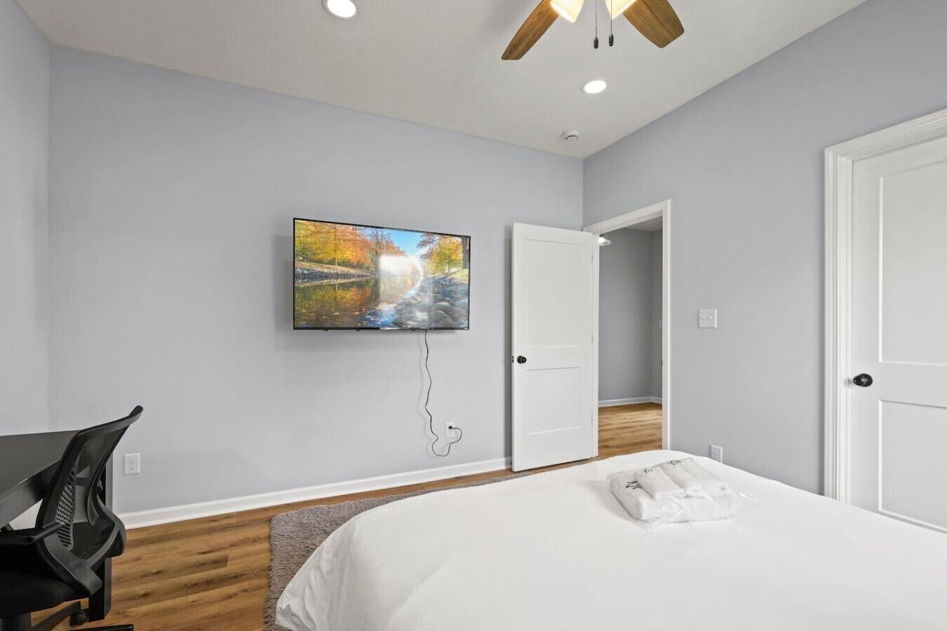 A bedroom with a bed, a desk, a chair, a painting, a ceiling fan, and a door leading to another room.