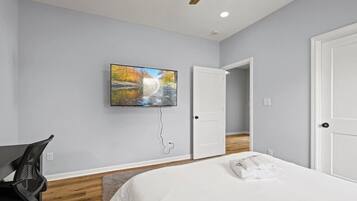 A bedroom with a bed, a desk, a chair, a painting, a ceiling fan, and a door leading to another room.