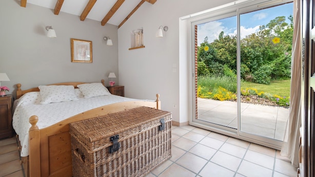Een slaapkamer met een bed, een rieten kist, een nachtkastje, een lamp en uitzicht op een tuin via een glazen schuifdeur.