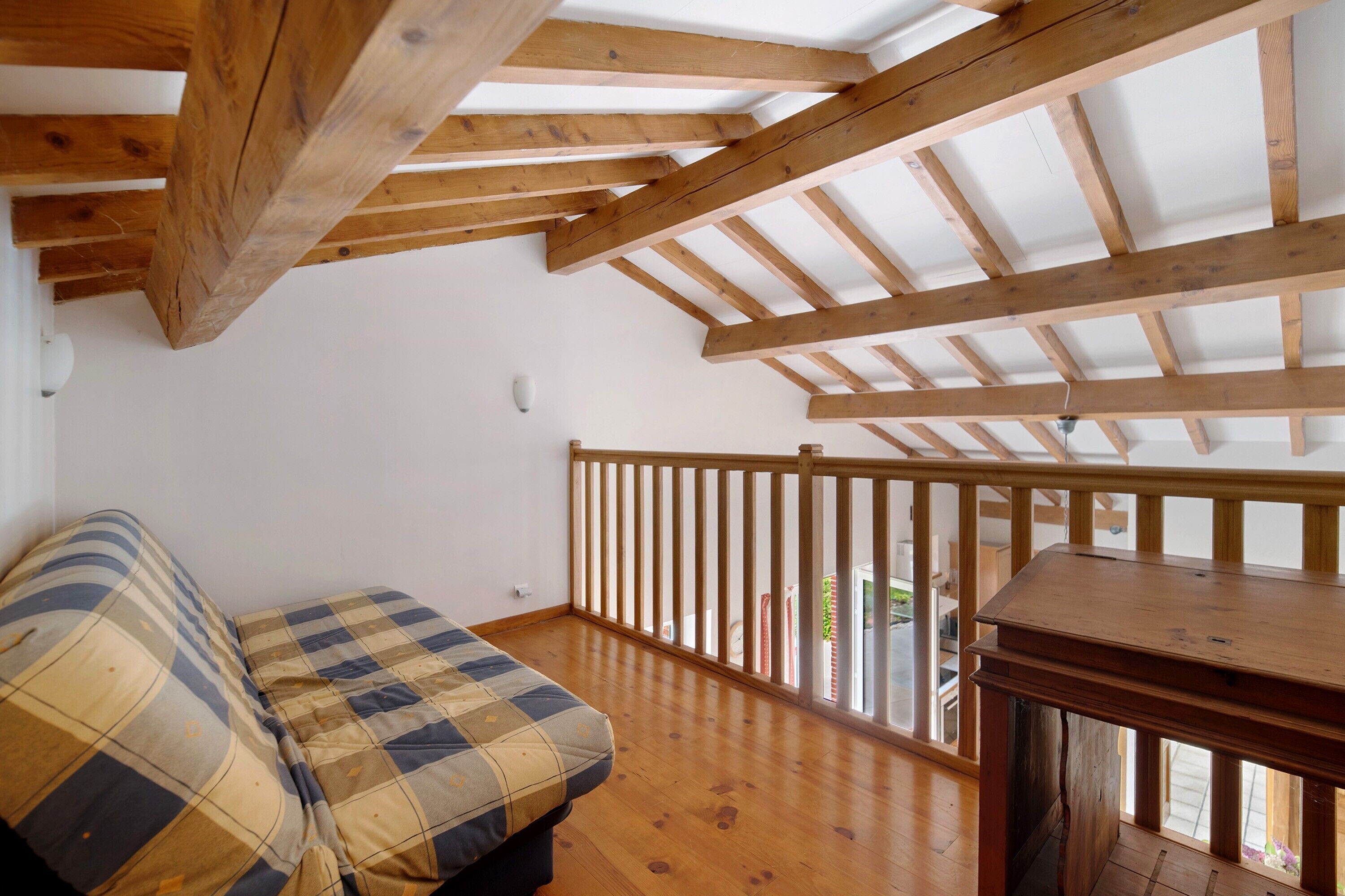 Een kamer met een houten plafond, een bank met een geruit kussen, een houten tafel en een balkon met een hekwerk.