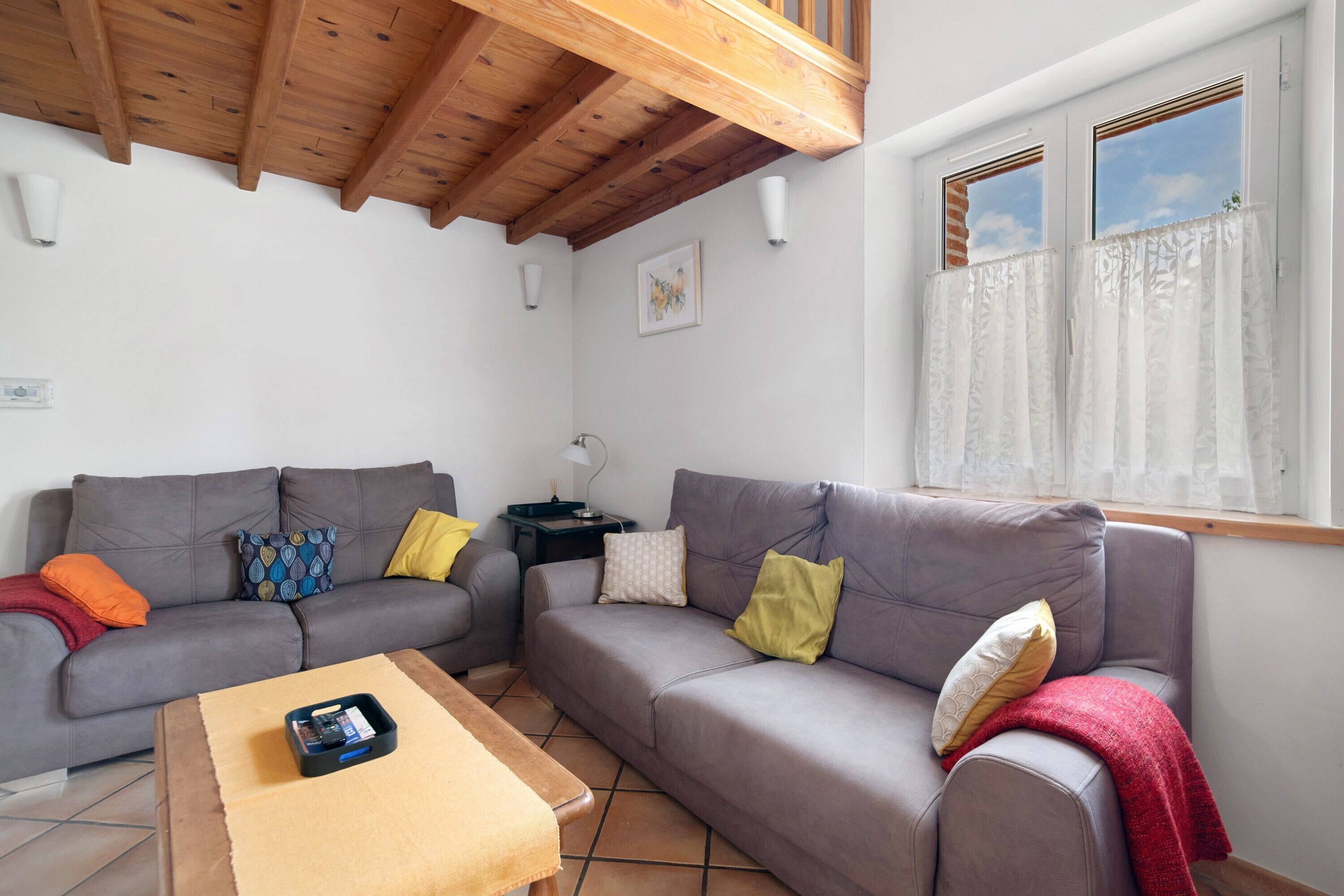 Una sala de estar con dos sofás grises, una mesa de centro, techo de madera, una ventana con cortinas y una pequeña mesa con una lámpara.