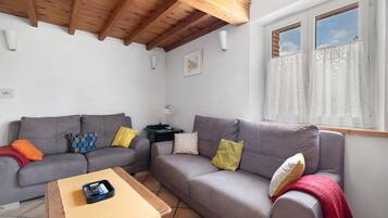Una sala de estar con dos sofás grises, una mesa de centro, techo de madera, una ventana con cortinas y una pequeña mesa con una lámpara.