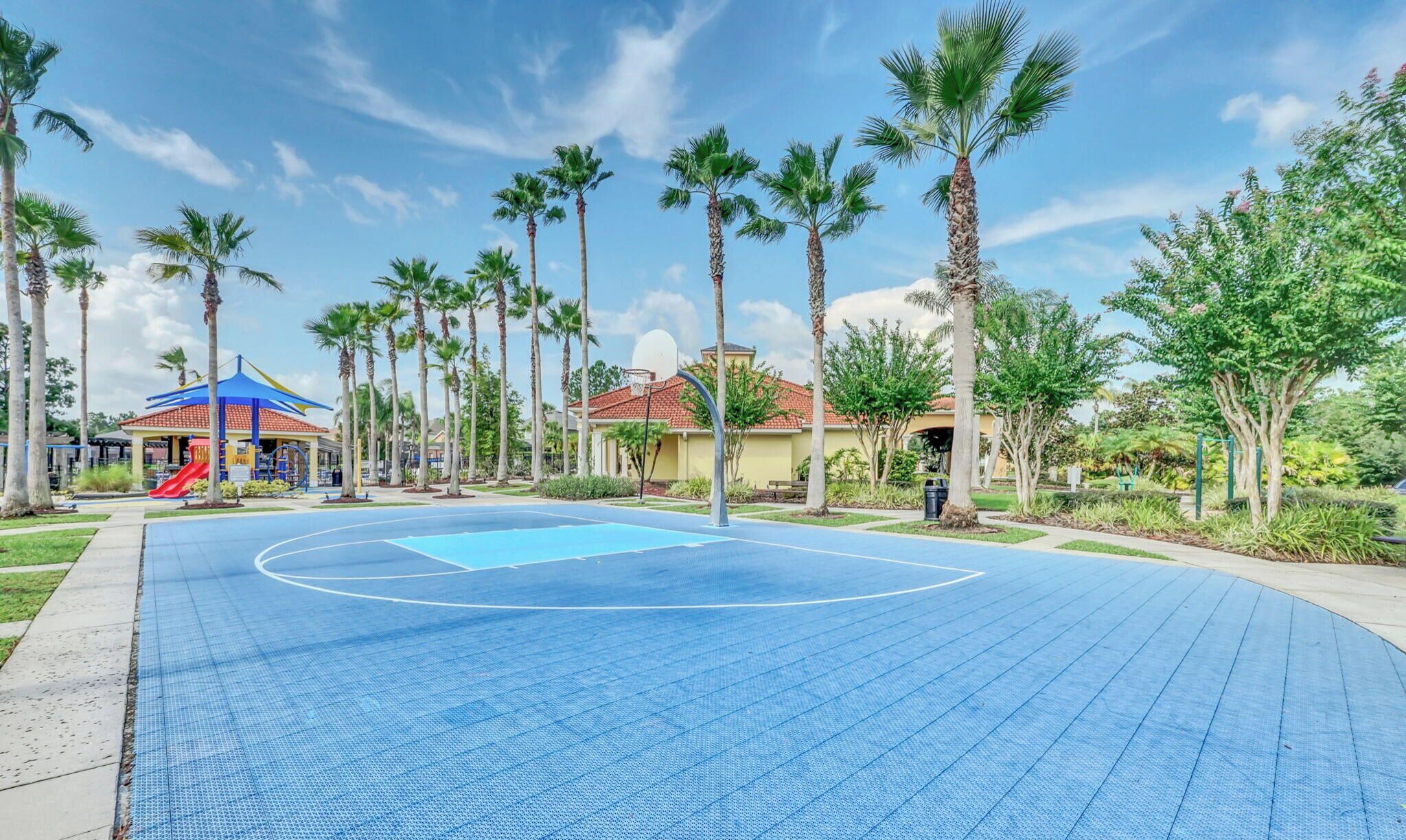 A basketball court with a blue surface, surrounded by palm trees and a playground with a red slide.
