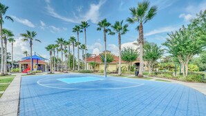 Un terrain de basket avec une surface bleue, entouré de palmiers et un espace de jeux avec un toboggan rouge.