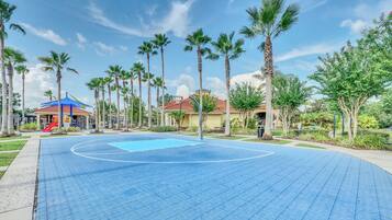A basketball court with a blue surface, surrounded by palm trees and a playground with a red slide.