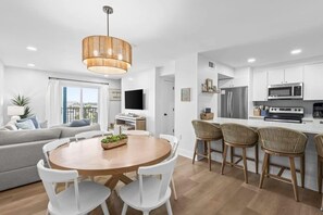 Eine moderne Küche mit weißen Schränken, ein Essbereich mit rundem Tisch und weißen Stühlen sowie eine Wohnbereich mit Sofa und Fernseher.