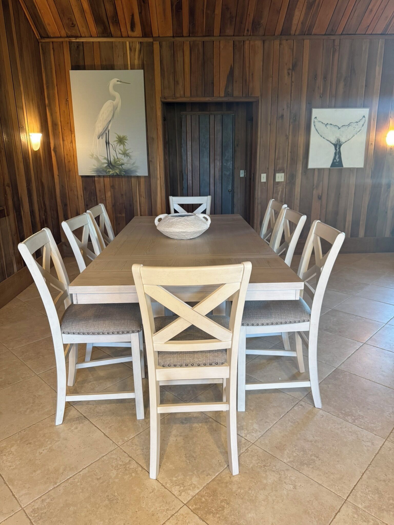 A dining area with a wooden table and white chairs, a wooden wall with a door, and two framed artworks.
