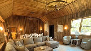 Ein geräumiges Wohnzimmer mit einer großen Eckcouch, einem Couchtisch und einem Teppich. Zwei Sessel und ein kleiner Tisch mit einer Lampe stehen ebenfalls im Raum. Die Wände sind aus Holz und die hohe Decke wird von einer Pendelleuchte erhellt.