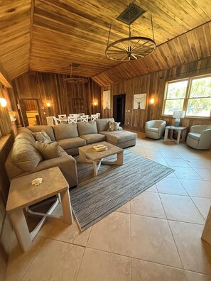 Ein geräumiges Wohnzimmer mit einer großen Eckcouch, einem Couchtisch und einem Teppich. Zwei Sessel und ein kleiner Tisch mit einer Lampe stehen ebenfalls im Raum. Die Wände sind aus Holz und die hohe Decke wird von einer Pendelleuchte erhellt.