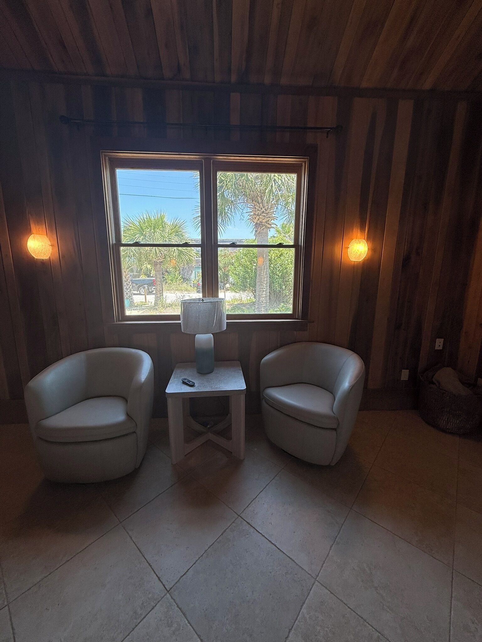 Una habitación con paredes de madera, dos sillones blancos, una mesita con una lámpara y una ventana con vista a palmeras.