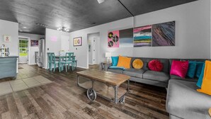 Una sala de estar moderna con un sofá gris, cojines coloridos, una mesa de centro de madera y un área de comedor con sillas color turquesa.