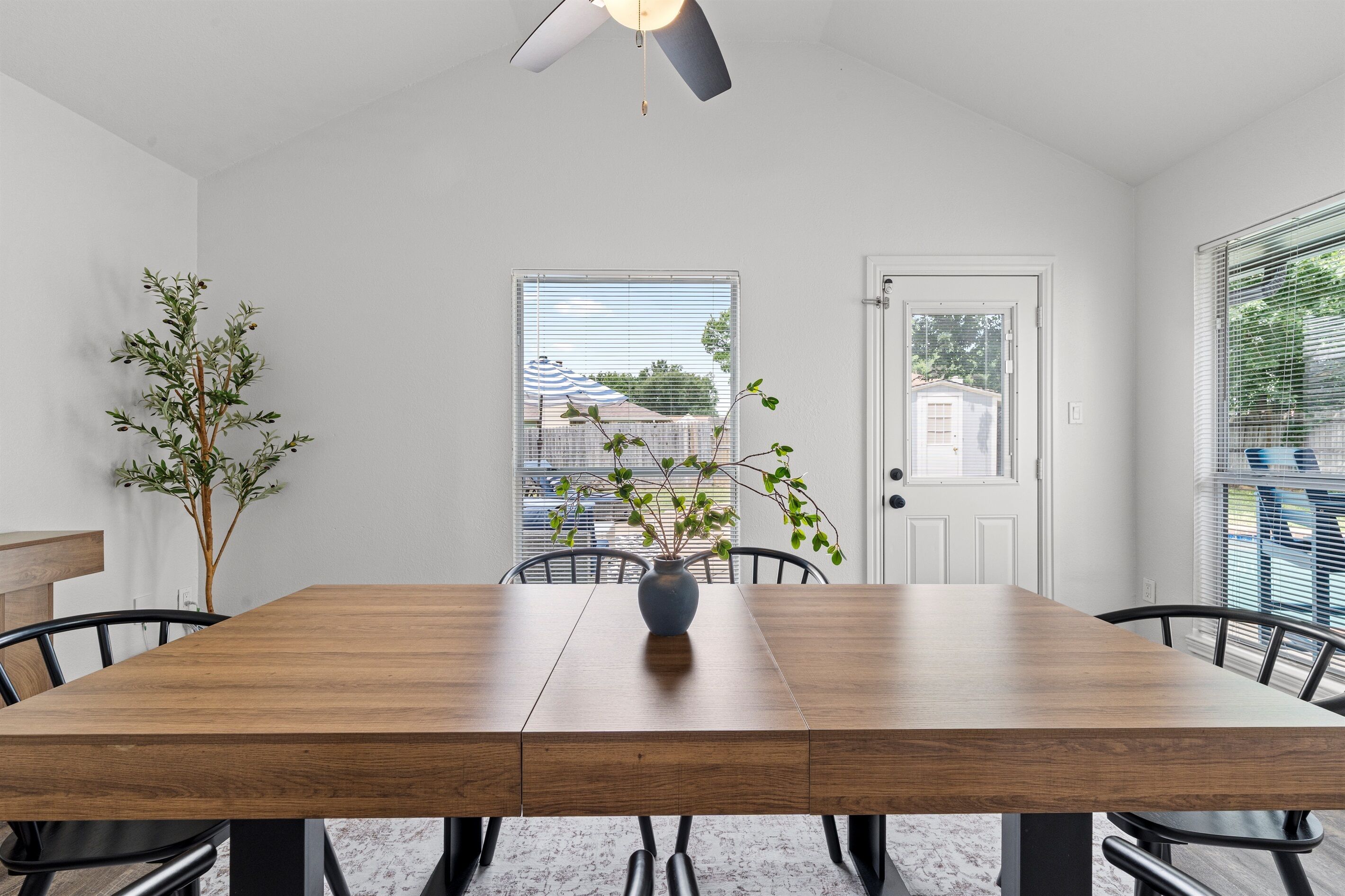 A dining area with a wooden table, chairs, a vase with a plant, a ceiling fan, and a view of the outside through a window.