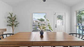 A dining area with a wooden table, chairs, a vase with a plant, a ceiling fan, and a view of the outside through a window.