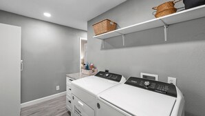 A laundry room with two stacked washers and dryers, a shelf with baskets, and a countertop with cleaning supplies. - Near AT&T Stadium | 3/2 Home with Boho Vibes (Arlington)