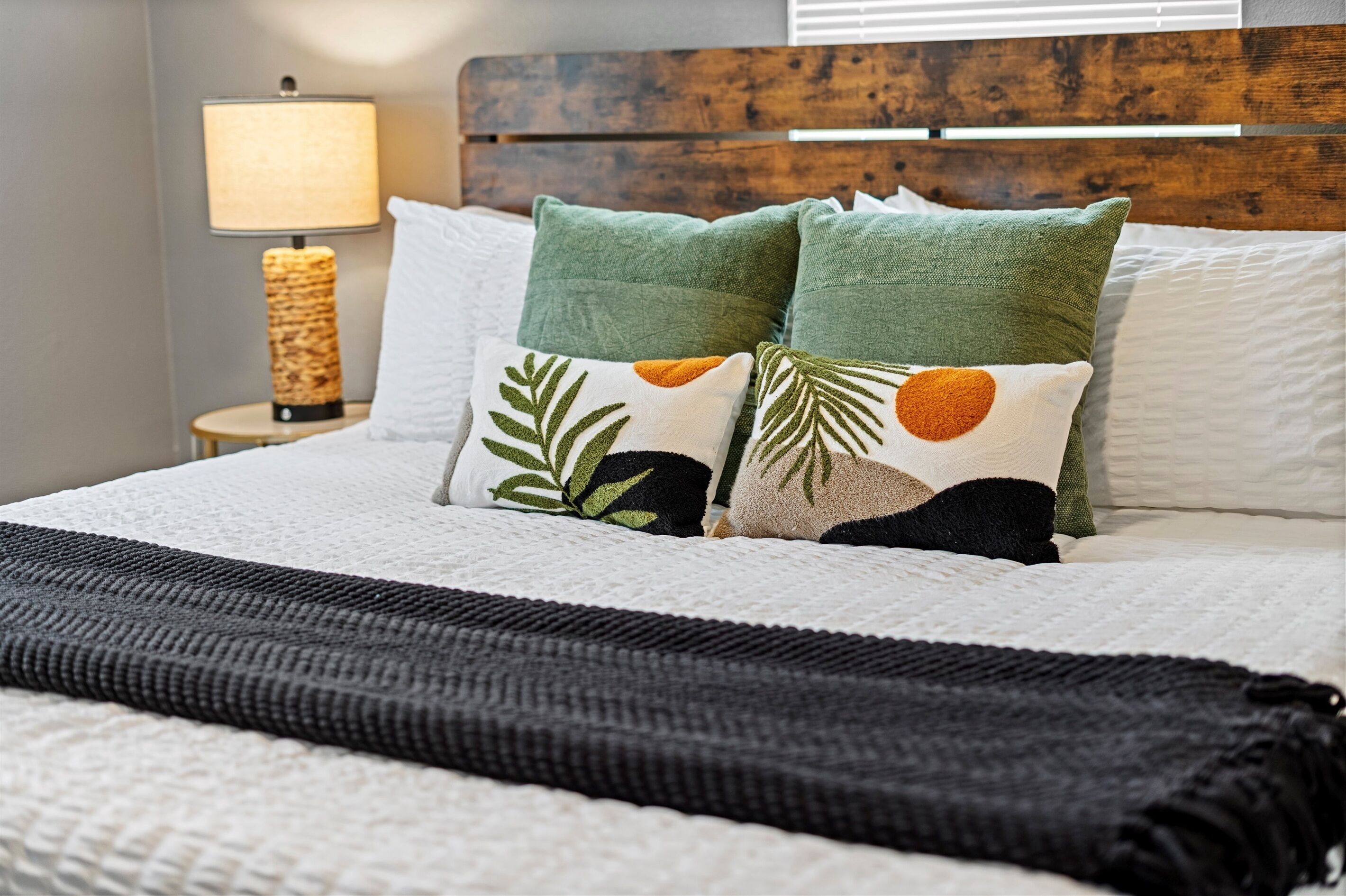 A neatly made bed with a white comforter, two green pillows with leaf patterns, a black throw blanket, and a wooden headboard.