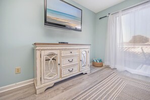 A room with a TV mounted on the wall, a white cabinet, a wooden floor, and a large window with sheer curtains. - The 312 at 69th Ave: Poolview Retreat (Myrtle Beach)