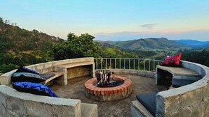 Uma área de estar circular ao ar livre com lareira, rodeada por paredes de pedra e guarda-corpo de metal, com vista para uma paisagem montanhosa.