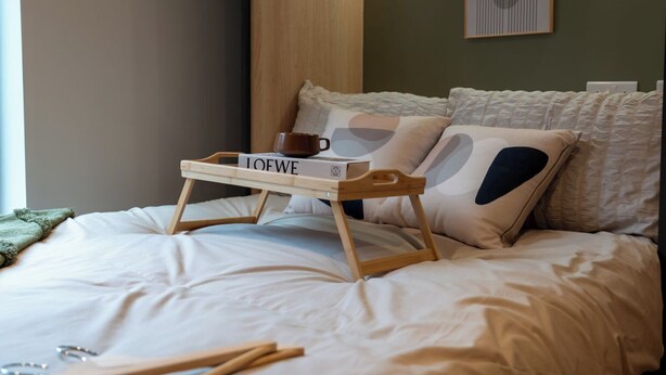 Een net opgemaakt bed met een houten dienblad waarop een boek en een kopje staan, een kussen met een opvallend design, en een abstract kunstwerk in een lijst aan de muur.