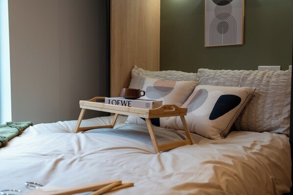 Ein ordentlich bezogenes Bett mit einem hölzernen Tablett, das ein Buch und eine Tasse trägt, ein Kissen mit einem besonderen Design und ein gerahmtes abstraktes Kunstwerk an der Wand.