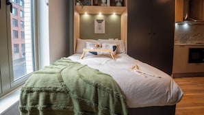 Een slaapkamer met een bed, een groene deken, een houten plank boven het bed en uitzicht op de straat door een raam.